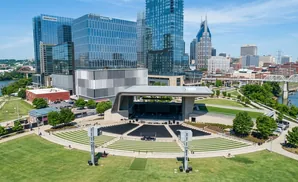 Aerial view of a modern amphitheater with tiered seating and a stage, backed by a city skyline with glass skyscrapers and a river.