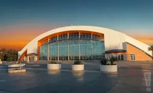 Modern arena with a curved roof and large glass facade, set against a sunset sky.