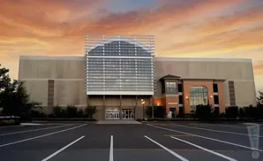 A large building with a glass facade and a tan exterior, set against a colorful sunset sky, with a parking lot in the foreground.