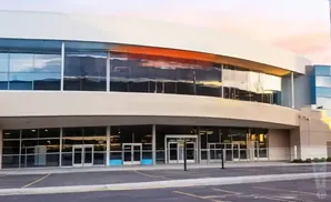 Exterior of a modern building with large glass windows reflecting a sunset, with parking lot in foreground.