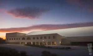 A long, tan building with arched windows and a curved roof is set against a sunset sky with pink and orange clouds.