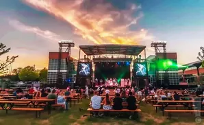 Outdoor concert at dusk with a band on stage, screens on the sides, and an audience seated at picnic tables.