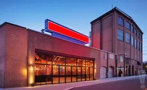 Exterior of a brick building with large glass doors, string lights inside, and a red billboard on top.