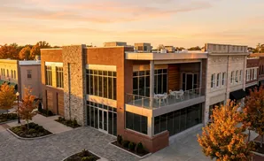 Modern brick building with stone accents, glass windows, and a balcony overlooking a street with autumn trees.