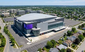 Aerial view of a large modern arena with a curved roof and purple digital screens, surrounded by parking lots and a suburban neighborhood.