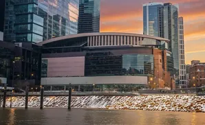 Modern arena with glass facade and tall skyscrapers behind, at sunset with snow-dusted riverbank.