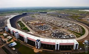 Aerial view of a large oval racetrack with grandstands, a parking lot full of vehicles, and surrounding fields.