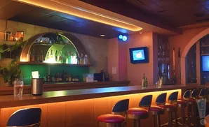 Bar with illuminated counter and red stools, bottle-lined shelves, and a television screen.