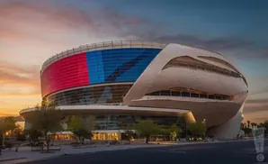 A modern arena with a curved facade, lit up with red and blue panels against a sunset sky.