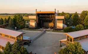 An outdoor concert stage with large screens, seating, and surrounding trees at sunset.