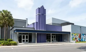 A modern building with purple accents and a colorful mural, flanked by palm trees and a street.