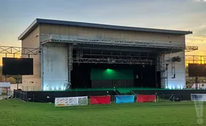 Outdoor concert stage with large screens and banners in front of a green field at sunset.