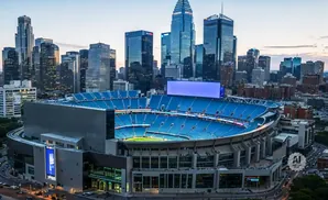 Sports stadium with blue seats and scoreboard in front of a city skyline at dusk.