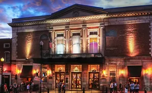 Exterior of an ornate brick building with large windows and classical columns, lit up at dusk.