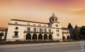 A cream-colored building with a domed tower and arched walkways against a warm, orange sky.