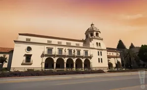 A large, Spanish Colonial-style building with a dome, arches, and balconies under a warm, orange sky.