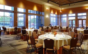 A banquet hall with round tables set for an event, large windows, and warm lighting.