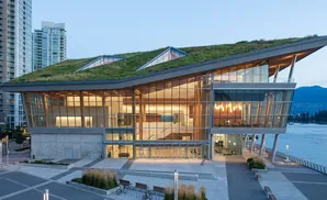 Modern building with a green roof by the water, featuring large glass windows and wooden beams.