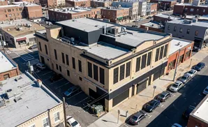 Aerial view of a tan brick building with dark windows, facing a street with parked cars and other buildings.