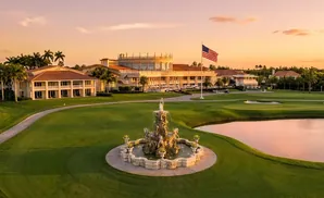 Golf course at sunset with a fountain, clubhouse, and palm trees.