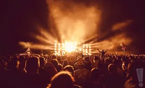 A crowd cheers at a concert, silhouetted against bright stage lights and smoke.