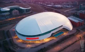 Aerial view of a large domed stadium with a white roof and a red sign, surrounded by modern buildings and city lights.