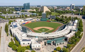 Aerial view of a baseball stadium in a city with tall buildings, a pyramid structure, and a golden bridge.