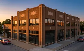 Art deco building with large windows and brick facade, parked vintage cars on street.
