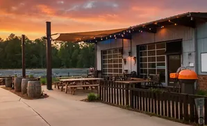 Outdoor seating area with picnic tables, barrels, and a grill under a sunset sky.