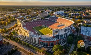 Aerial view of a large, open-air stadium with red seating, surrounded by trees and buildings.