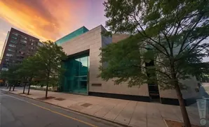 Modern building with large glass windows and stone facade, surrounded by trees, under a colorful sunset sky.