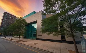 Modern building with large glass windows and beige stone facade, framed by trees and a vibrant sunset sky.