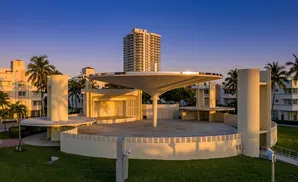 An outdoor stage with a large, umbrella-like roof and surrounding buildings and palm trees at sunset.