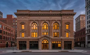 An ornate brick building with arched windows glows warmly at dusk, surrounded by other historic city structures.