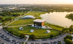 Aerial view of an outdoor amphitheater stage, lake, and parking lot at sunset.