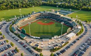 Aerial view of a baseball stadium, its field, grandstands, and surrounding parking lots filled with cars.