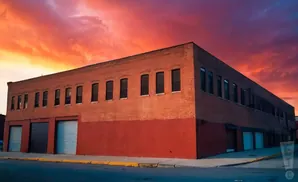 Brick building with red lower half and many windows under a dramatic orange and purple sunset sky.