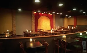 Intimate comedy club with tables and chairs, a stage with red curtains, and soft lighting.