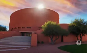 A large, round, terracotta building with a grand staircase leading to the entrance, framed by trees and a vibrant sunset sky.