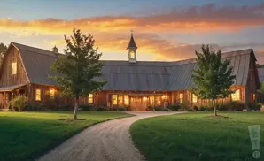 A long wooden barn with a metal roof at sunset, with a curved gravel driveway and green lawn.