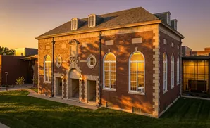 Brick building with arched windows and dormers bathed in golden sunset light, grass in foreground.