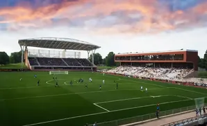 Soccer game at a stadium with two grandstands, one covered, under a colorful sunset sky.