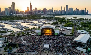 An aerial view of a concert venue with a large crowd gathered for a performance against a city skyline at sunset.