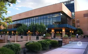 A modern building with large windows and a tan facade, with trees and outdoor seating.
