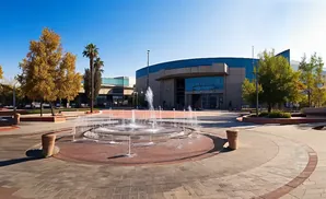 A circular fountain sprays water in front of a modern building with large windows, surrounded by trees and paved walkways.