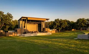 Outdoor amphitheater stage with musical instruments and speakers, set against a backdrop of trees under a clear blue sky.