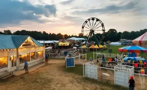 An outdoor fairground with rides, games, and food stands at sunset, with a Ferris wheel and a carousel.