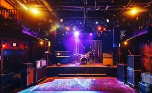 A concert venue stage with a drum kit, piano, amplifiers, and a rug on the floor, lit with purple and orange lights.