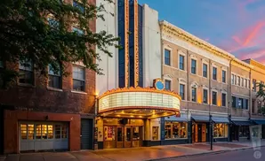 A vintage theater marquee illuminated at dusk on a city street, with brick buildings and shops lining the sidewalk.