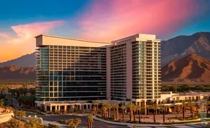 A modern hotel with glass walls stands before a mountain range under a vibrant sunset sky.